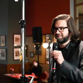 Andreas Scherffig an einem Mikrofon im Kaffeehaus. Im Hintergrund sitzt eine Person an einem Tisch.