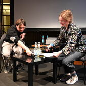 Elias Hirschl und Hanna Hamel sitzen an einem Tisch. Elias Hirschl greift zum Mikrofon, das auf dem Tisch liegt, Hanna Hamel lacht.