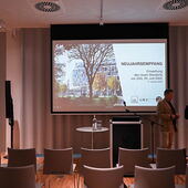 Auf einer Leinwand wird der Neujahrsempfang zur Einweihung des neuen Standorts von ZAS, ZfL und GWZ angekündigt. Davor stehen Stuhlreihen, rechts vor der Leinwand stehen zwei Personen.