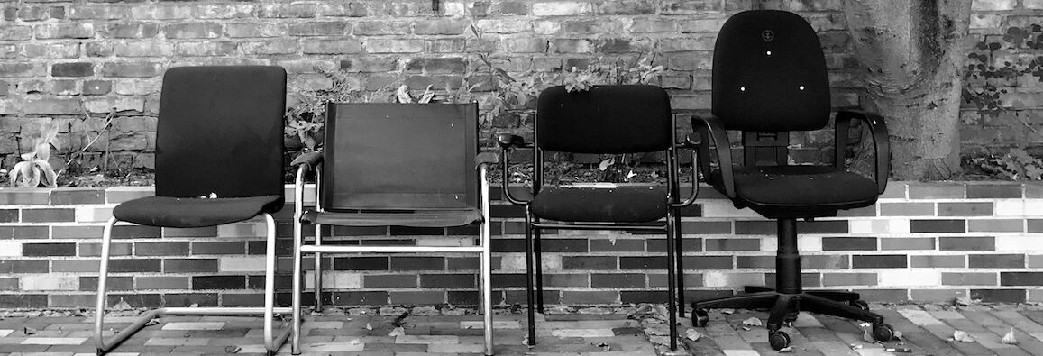 Black and white photo: Four different chairs are standing in front of a brick wall, from left to right: a curved chair without armrests, a cantilever chair, a chair with the backrest cut out at the bottom and armrests, an office chair with castors.
