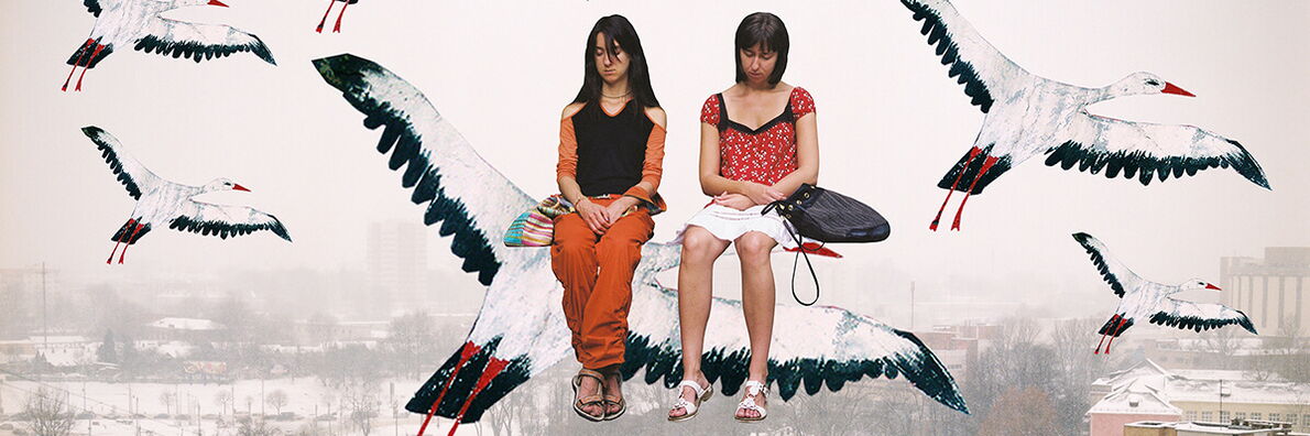 Two girls are sitting on a stork that is flying in a flock over a winter city shrouded in fog, with block buildings in the background.