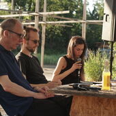 Cord Riechelmann, Moritz Gansen and Tabea Hertzog sit at a table made of chipboard on the open-air stage at Floating Berlin. Tabea Hertzog reads from a text, Cord Riechelmann and Moritz Gansen listen to her.