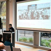 Eva Geulen steht am Pult und spricht ins Mikrofon. Hinter ihr auf der Leinwand ist ein Klassenfoto mit Duala Misipo zu sehen.