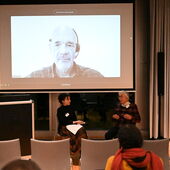 Sandra Folie und Ulfa von den Steinen sitzen auf Stühlen neben dem Rednerpult und unterhalten sich. Hinter ihnen auf der Leinwand ist Jürg Schneider per Zoom dazugeschaltet.