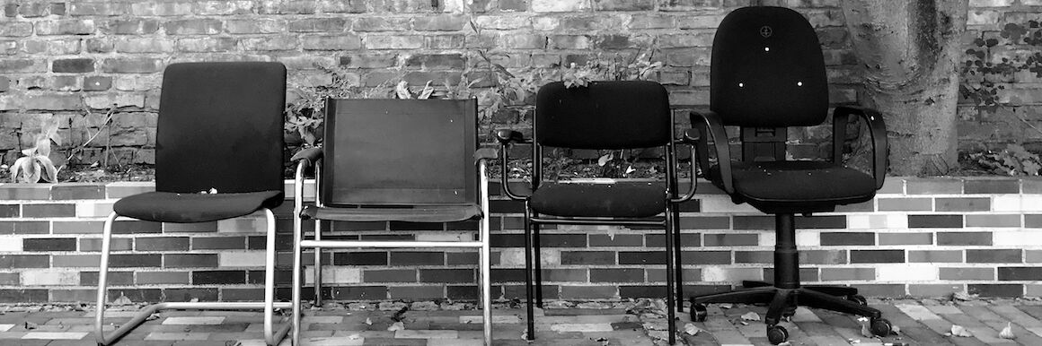 Black and white photo: Four different chairs are standing in front of a brick wall, from left to right: a curved chair without armrests, a cantilever chair, a chair with the backrest cut out at the bottom and armrests, an office chair with castors.