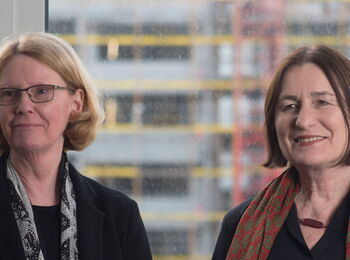Franziska Thun-Hohenstein and Irina Scherbakowa stand side by side in front of a window, behind which a construction site can be seen, and smile at the camera.