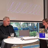 Alhierd Bacharevič und Tina Wünschmann sitzen vor der Leinwand und unterhalten sich, vor ihnen liegen Papiere und Bücher.