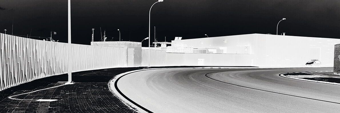 Negative image of a sharp street curve lined with lanterns. In the upper right, the outline of a car can be seen in front of an overlit building.