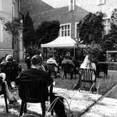 In einem kleinen Garten sitzen Menschen auf Gartenstühlen. Sie sind von hinten zu sehen und schauen auf ein kleines Zelt mit einer Bühne.