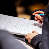 Ein DIN-A4-Heft mit handschriftlichen Notizen wird von einer linken Hand gehalten, in der rechten Hand der Person ist ein blauer Kugelschreiber zu sehen.