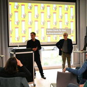 Christoph Hesse, Michael Bienert and Dirk Naguschewski stand in front of a screen on which the same image of an advertising pillar with two people hiding behind it can be seen several times on a yellow background.
