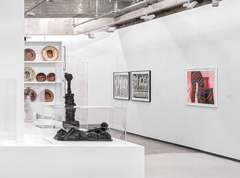 An exhibition space featuring sculptures, ceramic plates, woodcuts, and colorful paintings.