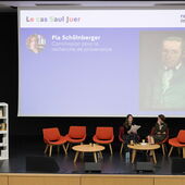 Zwei Personen sitzen auf dem Podium und sprechen miteinander. Auf der Leinwand hinter ihnen steht »Le cas Saul Juer«, darunter sind ein kleines Porträt von Pia Schölnberger und rechts davon eine Porträtmalerei zu sehen.