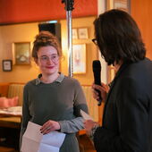 Hanna Hamel and Andreas Scherffig are standing in front of a podium. Scherffig can be seen from behind, holding a microphone and talking.