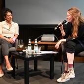 Elisa Aseva und Hanna Hamel sitzen auf einem Podium vor einer Leinwand. Hanna Hamel hält ein Mikrofon in der Hand.