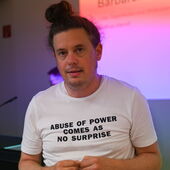 Matthias Meindl stands in front of a wall onto which intense purple and red are projected. He’s wearing a white T-shirt that says “Abuse of Power Comes as No Surprise.”"
