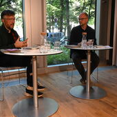 Lukas Bärfuss und Stefan Willer sitzen nebeneinander an weißen Bistrotischen und unterhalten sich.