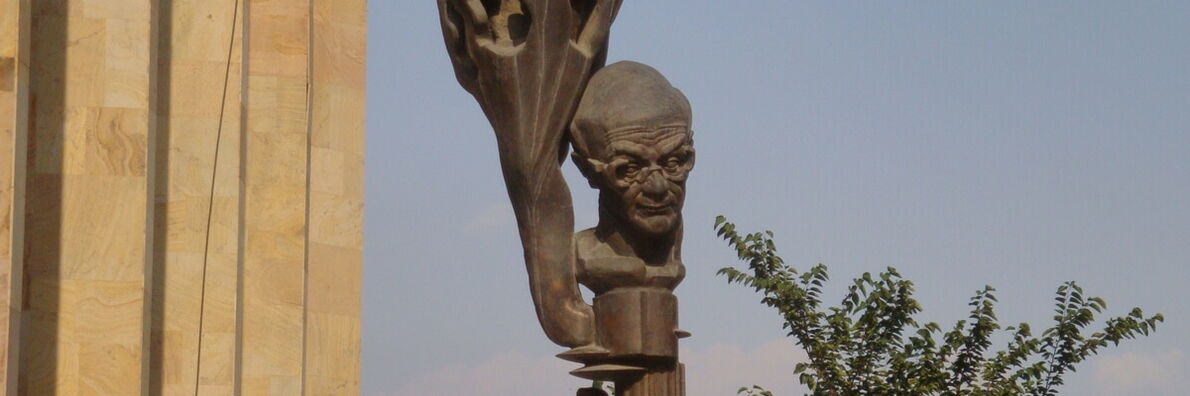 A bronze statue of the philosopher Merab Mamardashvili against a blue sky. Branches extend into the frame on the right.