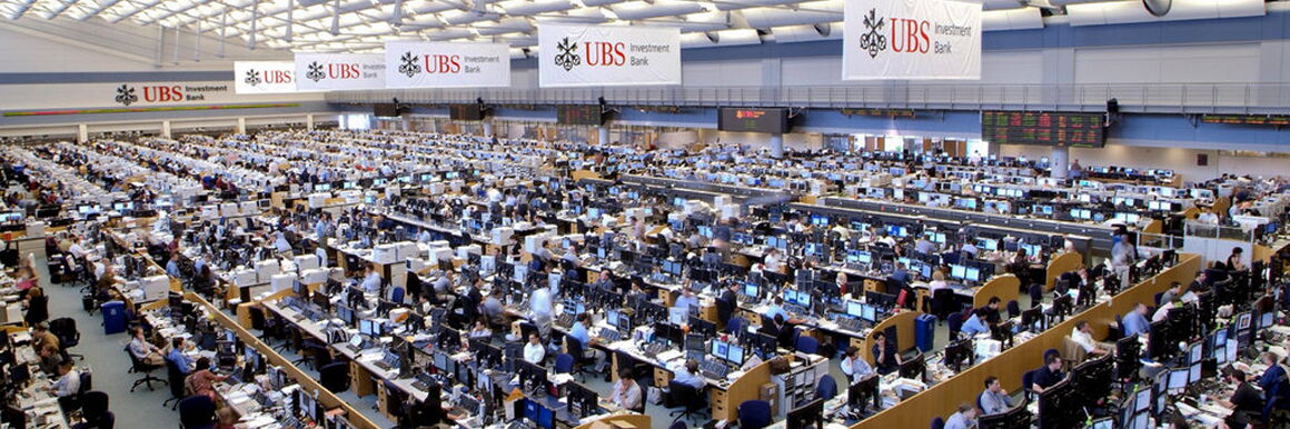 Photo of the UBS trading floor from above. A great many people are sitting in a large hall at computers set up in row-like blocks. Above them hang several white and red banners of UBS.