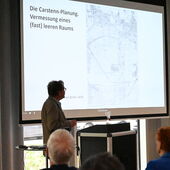 Andreas Ludwig stands at the lectern and turns to the screen behind him, on which a map of the city and text about the Carstenn figure can be seen.