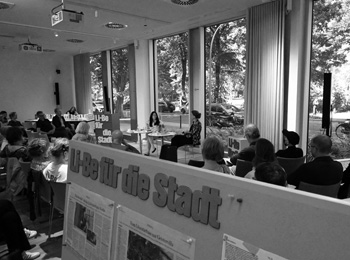 A full event room. At the front, a board with newspaper cuttings reads “Li-Be für die Stadt”.